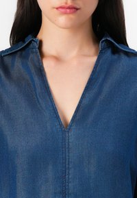 Denimskjorta med djup V-ringning, kort krage och subtil glans. Den har en strukturerad passform med synliga sömdetaljer.