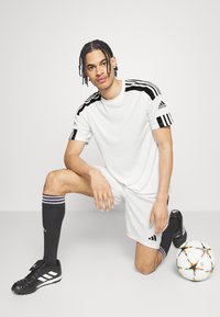 Vit atletisk tröja och shorts med svarta detaljer, svarta strumpor med ränder, svarta fotbollsskor och en flerfärgad fotboll på en ljus bakgrund.