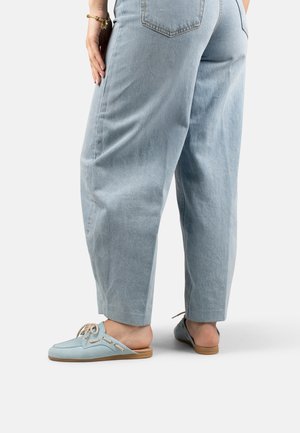 Lichtblauwe denim jeans gedragen met lichtblauwe slip-on loafers met veters, van achteren getoond op een effen lichte achtergrond.
