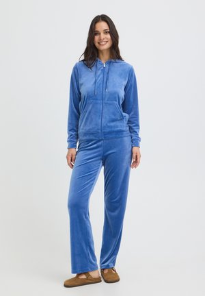 Žena sa usmieva, má na sebe modrú zamatovú mikinu na zips a zodpovedajúce nohavice s hnedými slip-on topánkami na jednofarebnom pozadí.