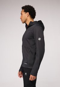Zwarte softshell jas met een rits aan de voorkant, een capuchon en zijzakken; beschikt over een logo op de linkermouw en een gladde, textuurrijke afwerking.