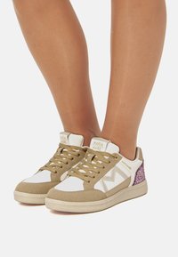 Zapatillas beige y blancas con un acabado de ante texturizado, que cuentan con un acento de brillo y una suela de goma. Diseño de cordones con una marca en la lengua.