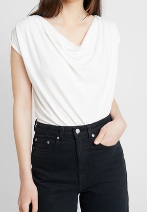 Kobieta w białej bluzce bez rękawów z drapowanym dekoltem, włożonej do czarnych, wysokich spodni jeansowych, z jedną ręką w kieszeni.