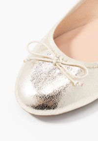 Metallisk guldig balettflats med rund tå, texturerad ovandel och en dekorativ rosett. Slät inredning, minimalistisk design.