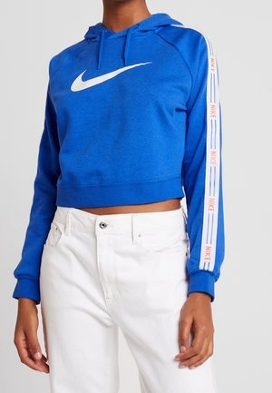 Sweat à capuche court bleu Nike avec logo swoosh blanc et bandes de marque sur les manches, porté avec un pantalon blanc taille haute.