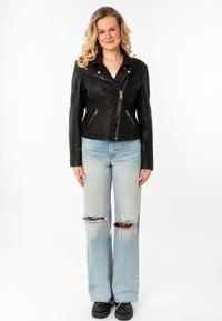 Zwarte leren motorjack met ritssluiting aan de voorkant, twee zijzakken en gouden hardware, gecombineerd met lichtblauwe ripped jeans en zwarte schoenen.