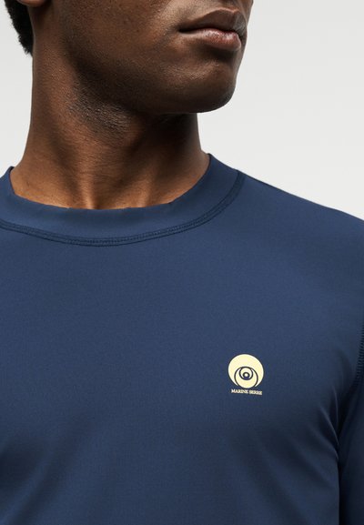 Κοντινό πλάνο ενός ατόμου που φοράει μπλε πουκάμισο με μικρό κίτρινο κυκλικό λογότυπο Marine Serre στο στήθος.