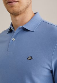 Polo de color azul claro con tejido texturizado. Presenta un logo bordado en negro en el lado izquierdo del pecho y un cuello puntiagudo con dos botones.