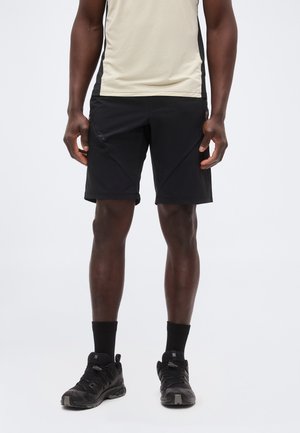 Mężczyzna w beżowej koszulce bez rękawów, czarnych sportowych szortach, czarnych skarpetkach i czarnych butach do biegania stoi na białym tle.