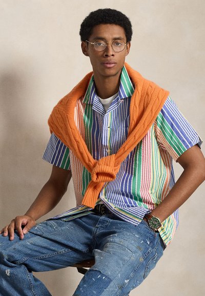 Πουκάμισο με πολύχρωμες ρίγες και κοντά μανίκια, με πορτοκαλί πουλόβερ περασμένο στους ώμους, συνδυασμένο με μπλε τζιν με φθορές και πράσινο ρολόι.