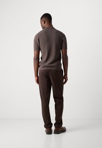 Suéter de punto marrón de manga corta, combinado con pantalones marrones ajustados. El modelo está de espaldas, mostrando la textura del tejido del suéter.