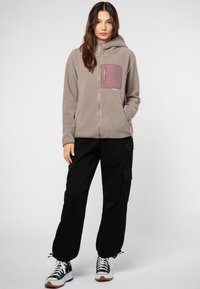 Fleece jack in lichtgrijs met een paarse ritszak op de borst, een rits aan de voorkant en een capuchon. Zwarte cargo broek en sneakers maken de outfit compleet.