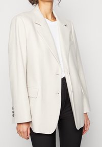 Blazer oversize grigio chiaro con due bottoni, tasche frontali e colletto a rever. Il tessuto ha una texture liscia, abbinato a una maglia bianca e pantaloni neri.