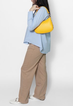 Borsa a mano gialla in pelle, con forma arrotondata, hardware in argento e superficie liscia, indossata sopra una camicia azzurro chiaro con orlo curvo e pantaloni beige a righe.