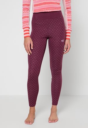 Frau trägt bordeauxrote Leggings mit Muster und ein langärmliges Oberteil mit pink-rotem Muster, steht barfuß vor einem schlichten Hintergrund.