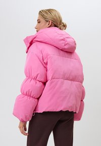 Žena s blond vlasmi zviazanými do drdolu, oblečená v jasne ružovej bundě a tmavých nohaviciach, otočená chrbtom k jednoliatej pozadiu.