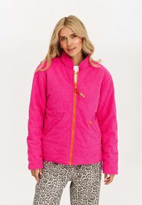 Jasnoróżowa kurtka z korduroju z wysokim kołnierzem, z pomarańczowym zamkiem błyskawicznym i kieszeniami bocznymi, noszona na spodniach w leoparda.