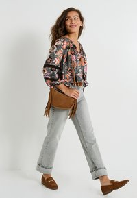Sac en suède marron avec des franges, blouse noire à motifs floraux colorés, jeans gris clair et mocassins marron. La tenue présente une coupe décontractée et un imprimé détaillé.