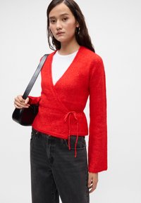 Červený svetr s dlouhými rukávy, texturovaná tkanina a vázání v oblasti pasu, v kombinaci s bílým tričkem a černými džíny.