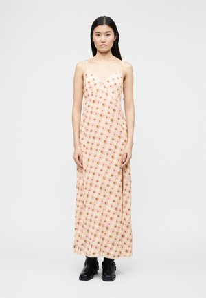 Femme aux longs cheveux noirs portant une robe nuisette rose à fleurs et des bottines noires, debout les bras détendus devant un fond uni.