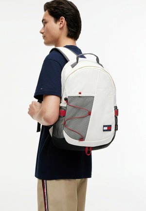 Młody mężczyzna w granatowej koszuli i beżowych spodniach, noszący biało-czarny plecak z siatkową kieszenią i czerwonymi detalami, stojący bokiem.