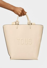 Borsa tote in pelle beige con finitura opaca, forma strutturata, logo "TOUS" impresso e manici annodati. Rifinita con bordi neri.