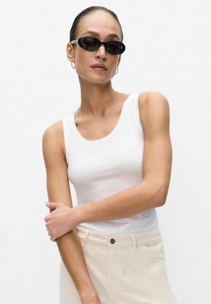 Femme portant des lunettes de soleil ovales noires, un haut blanc sans manches et un pantalon beige, posant les bras croisés devant un fond blanc uni.