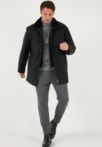 Černý kabát z vlněné směsi s vysokým límcem, zapínáním na zip a dvěma bočními kapsami; v kombinaci se šedými kalhotami na míru a černými koženými botami.