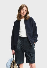 Mladá žena v námořnicky modré fleecové bundě, bílém topu a černých cargo šortkách, držící modrý a béžový batoh na jednobarevném pozadí.