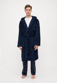 Marineblå, luksuriøs badekåpe med hette, frontknyting og sidelommer, brukt over hvit henley skjorte og marineblå rutete bukser.