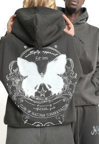 Dunkelgrauer Hoodie aus weichem Stoff, mit einem großen weißen Schmetterlingsgrafik und dekorativen Elementen sowie Text auf dem Rücken.