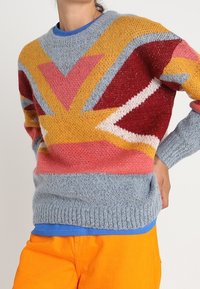 Pull tricoté avec un motif géométrique audacieux aux couleurs jaune, rouge, rose et gris. Poignets et ourlet côtelés, porté avec un pantalon orange.
