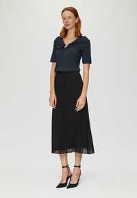 Zwarte plissé midi-rok gecombineerd met een donkere blauwe top met een sleutelgatdetail. Het model draagt zwarte enkelbandhakken. Gladde stofstructuur.