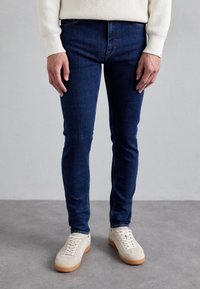 Granatowe, dopasowane jeansy z klasycznym wzorem pięciu kieszeni, zestawione z jasnobeżowymi trampkami o fakturowanej cholewce i gumowej podeszwie.