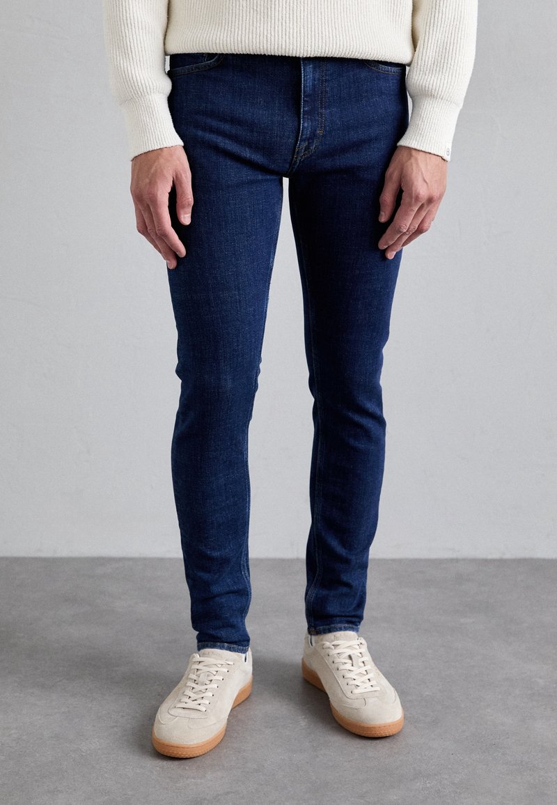 Tummansiniset slim-fit-farkut, joissa on klassinen viisipokkoinen muotoilu, yhdistetty vaaleanbeigeihin tennareihin, joissa on teksturoitu päällinen ja kumipohja.