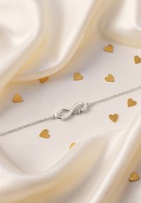 Braccialetto d'argento con un charm infinito dal design ondulato, decorato con piccole pietre trasparenti, su un tessuto satinato liscio con cuori dorati.