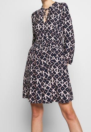 Woman wearing a navy blue dress with a pale pink geometric pattern, long sleeves, and a tied V-neckline, standing against a plain background.