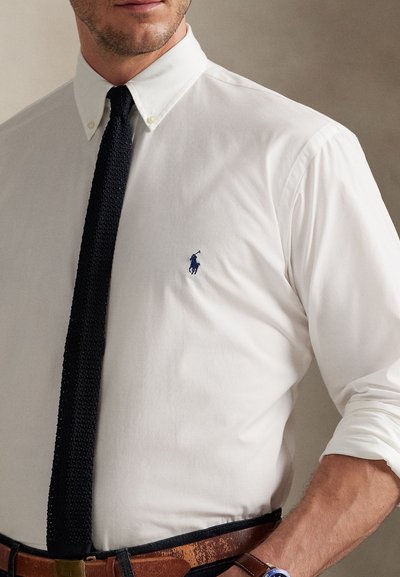 Man wearing white button-down shirt with navy embroidered logo, black knit tie, brown belt, and rolled-up sleeves.