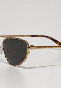 Ochelari de soare din metal aurit, cu lentile întunecate și un design unghiular. Brătările au un model țestoasă și logo-ul „Michael Kors” gravat.