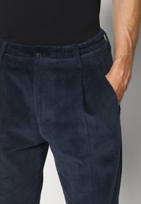 Homme portant un pantalon en velours côtelé bleu foncé avec poches latérales, main droite dans la poche, associé à une chemise noire ajustée.