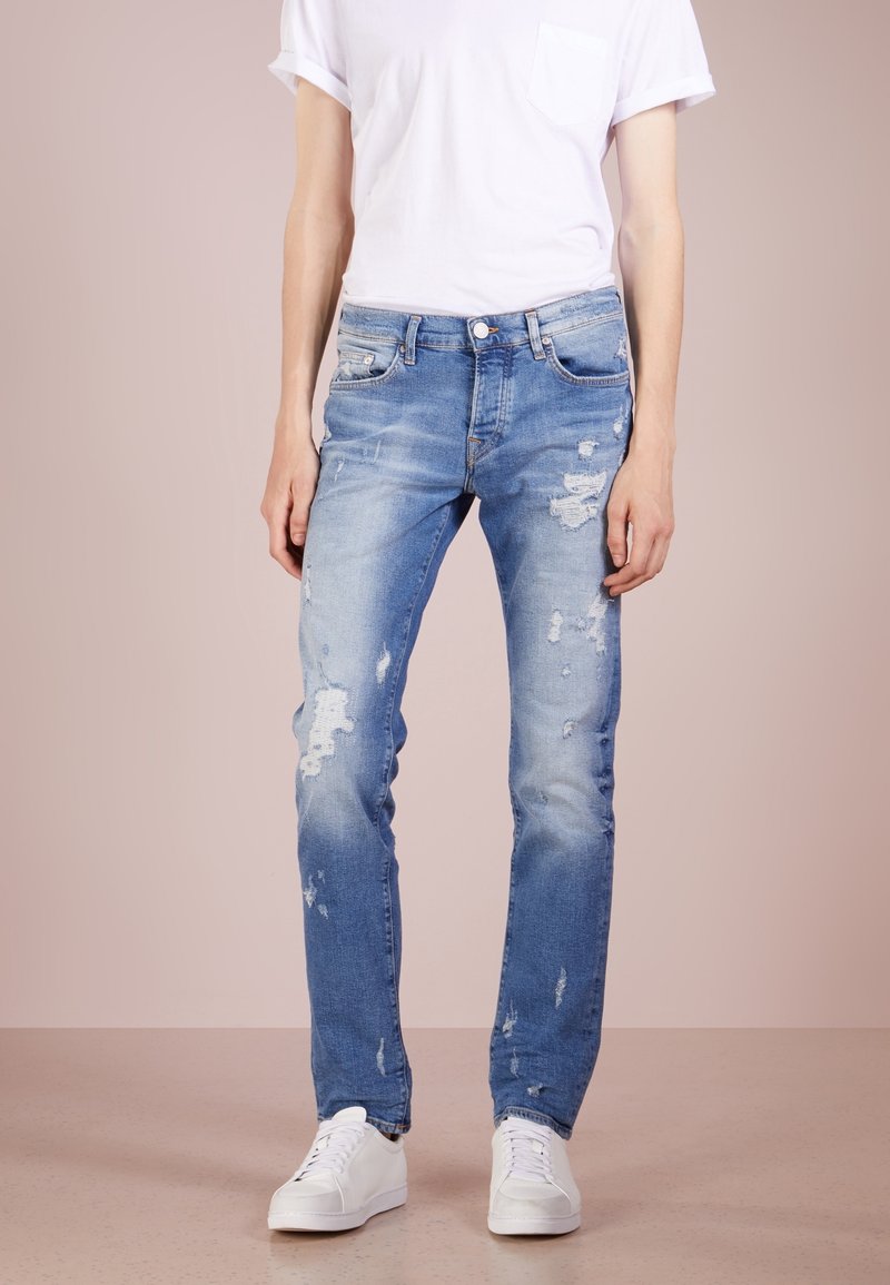 Ljusblå slitna denimjeans med hål och abrasioner, smal passform, ihop med en enkel vit t-shirt och vita sneakers.