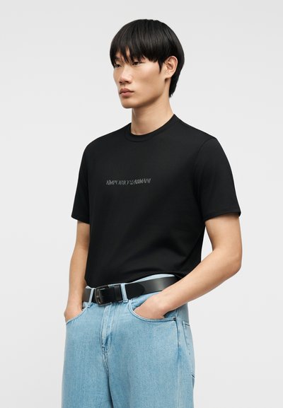 Νέος άνδρας με ίσια μαύρα μαλλιά φοράει μαύρο μπλουζάκι Emporio Armani, φορεμένο μέσα σε ανοιχτόχρωμο μπλε τζιν με μαύρη ζώνη, με τα χέρια στις τσέπες.