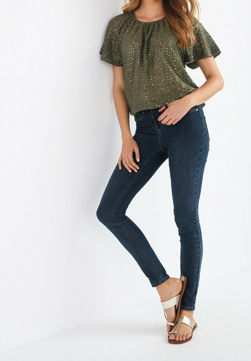 Femme portant un haut court vert olive avec des sequins, un jean skinny bleu foncé et des sandales beige à enfiler, debout sur fond blanc.