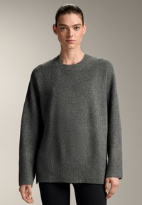Šedý pletený svetr s kulatým výstřihem, spadlými rameny a ribborovým lemem. Jemná struktura, mírně oversized střih, spojený s černými legínami.