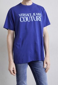 Camiseta de algodón azul con "VERSACE JEANS COUTURE" impreso en letras blancas en negrita. Mangas cortas y escote redondo. Combinada con jeans de mezclilla.
