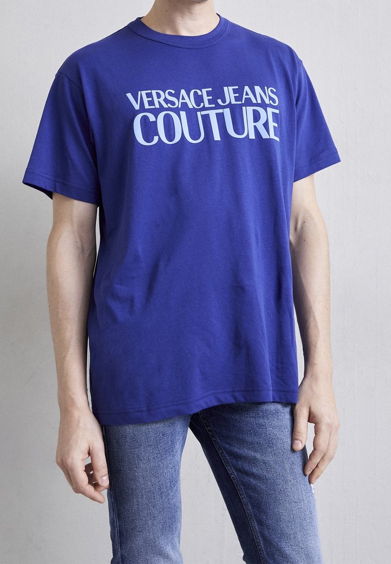 Camiseta de algodón azul con "VERSACE JEANS COUTURE" impreso en letras blancas en negrita. Mangas cortas y escote redondo. Combinada con jeans de mezclilla.