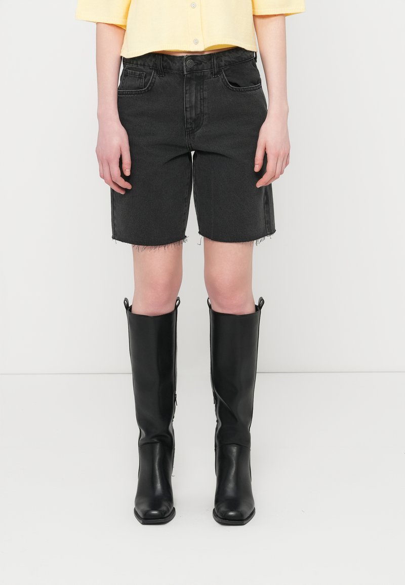 Μαύρα τζιν σορτς με φθαρμένη τελείωμα, συνδυασμένα με μαύρα δερμάτινα μποτάκια μέχρι το γόνατο. Το μοντέλο φοράει κίτρινο κοντό πουκάμισο με κουμπιά.