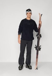 Schwarzer Fleece-Pullover, schwarze Skihose, graue Mütze und schwarze Skischuhe. Hält Skier mit Bindungen. Schlichter Hintergrund.