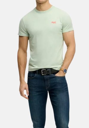 Hombre que lleva una camiseta ajustada de color verde claro, vaqueros azul oscuro, cinturón negro y reloj de pulsera con una mano en el bolsillo.