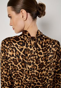 Jedwabna bluzka w panterkę z wysokim dekoltem, z tylnym wiązaniem i gładką teksturą, w odcieniach brązu i czerni.